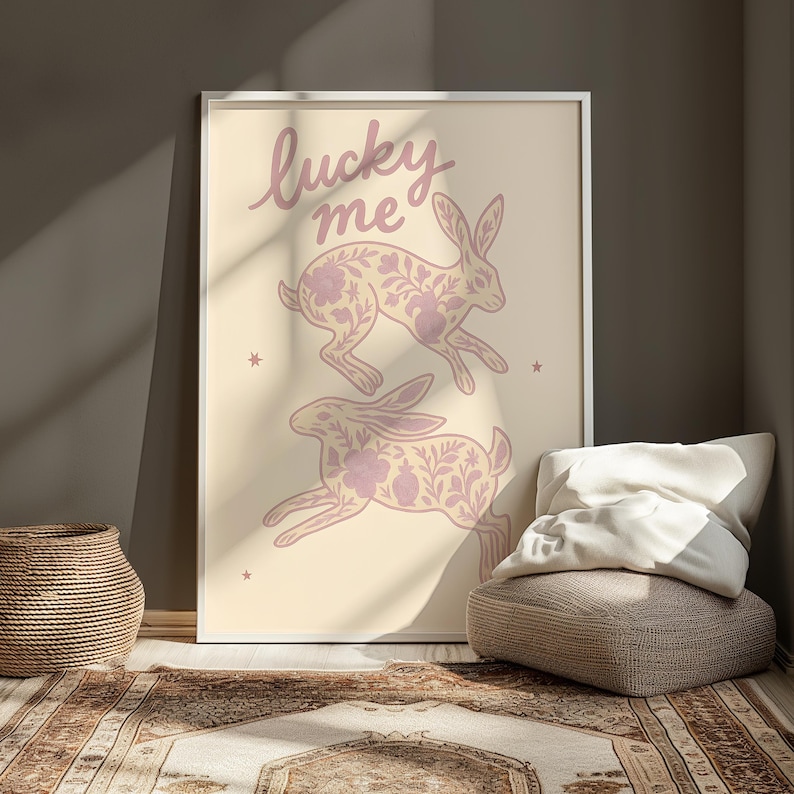 May include: A framed art print featuring two running rabbits with floral patterns in a soft purple hue. The words "lucky me" are written above the rabbits in a matching color. The background is a light beige.