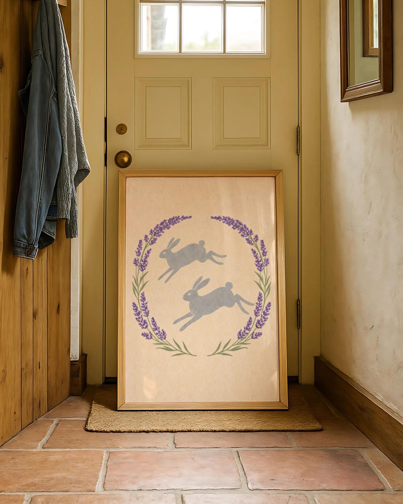 May include: A framed print showing two grey hares leaping within a lavender wreath. The artwork has a light beige background and is displayed in a natural wood frame. The print is leaning against a door.