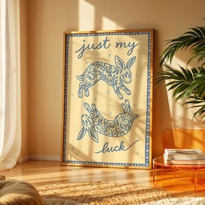 May include: A framed art print featuring two blue floral patterned rabbits on a cream background. The words "just my luck" are written in cursive. The print is in a wooden frame and is displayed in a well-lit room.