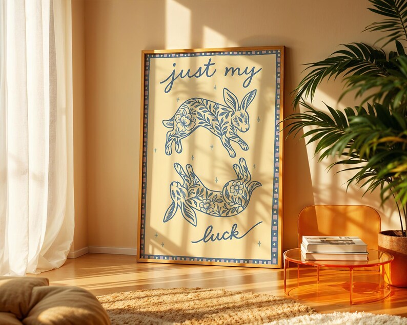 May include: A framed art print featuring two blue floral patterned rabbits on a cream background. The words "just my luck" are written in cursive. The print is in a wooden frame and is displayed in a well-lit room.