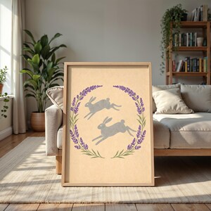 May include: Framed print with two grey hares leaping inside a lavender and green wreath. The artwork is set against a beige background, with a natural wood frame. The print is displayed in a living room setting.