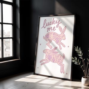 May include: Framed art print featuring two pink rabbits with floral patterns, and the words "lucky me" in cursive. The artwork is set against a white background with small star accents. The frame is black.