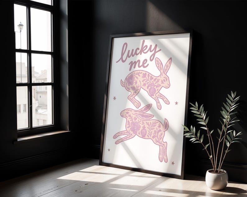 May include: Framed art print featuring two pink rabbits with floral patterns, and the words "lucky me" in cursive. The artwork is set against a white background with small star accents. The frame is black.