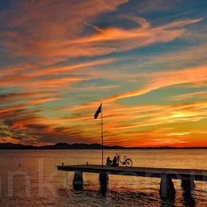 May include: A wooden pier extends into a calm body of water at sunset. The sky is a vibrant mix of orange, pink, and blue with wispy clouds. A lone figure sits on the pier with a bicycle.