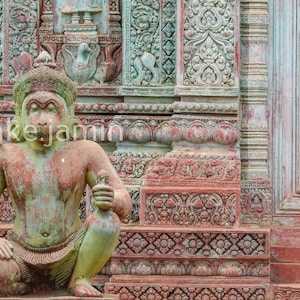 Puede incluir: Estatua sentada y desgastada con pátina verde y roja, sosteniendo una pequeña botella. La estatua está frente a una pared de piedra tallada con patrones florales y geométricos. La paleta de colores es apagada, con tonos de verde, rojo y gris.