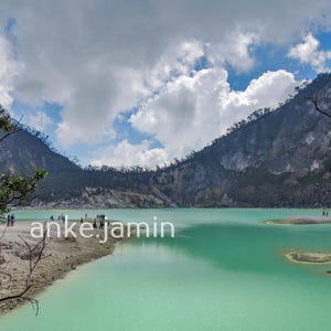 Puede incluir: Una vista panorámica de un lago volcánico de color turquesa rodeado de montañas rocosas escarpadas bajo un cielo azul brillante con nubes blancas. El texto anke.jamin es visible en letras blancas sobre el agua. Ramas desnudas enmarcan la escena.