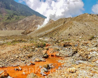 May include: A volcanic landscape featuring a stream of bright orange water flowing through a rocky, barren terrain. In the background, a mountain slope rises with a plume of white smoke or steam rising from the peak against a blue sky.