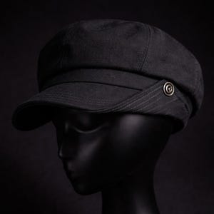 May include: A dark grey newsboy cap with a rounded crown and a short, stiff peak. The cap has a decorative button on the side and is made of a textured material. The hat is displayed on a black mannequin head.