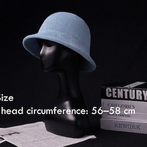 Op de afbeelding: Een lichtblauwe cloche hoed met een afgeronde kroon en een kleine rand, gepresenteerd op een zwarte mannequin hoofd. De hoed past op een hoofdomtrek van 56-58 cm. De achtergrond bevat boeken en een krant.