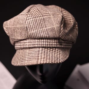 Peut inclure: Casquette de style gavroche à carreaux marron et blanc. La casquette a une couronne arrondie et une courte visière rigide. Le tissu présente un motif classique. La casquette est présentée sur une tête de mannequin noire sur fond noir.