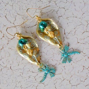 May include: A pair of gold-colored leaf-shaped earrings with teal accents. Each earring features a teal bead, small gold leaves, and a teal dragonfly charm. The earrings have gold-colored ear wires.
