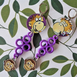 Puede incluir: Un par de pendientes colgantes con un diseño de mono de dibujos animados. Los pendientes tienen una cadena púrpura de círculos y un dije de cara de mono. La parte superior de los pendientes es redonda con una cara de mono sobre un fondo púrpura.