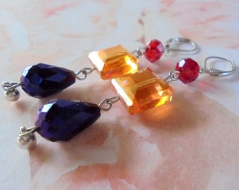 May include: A pair of dangle earrings with a teardrop-shaped purple crystal, a square orange crystal, and a round red crystal. The earrings are made with silver-toned metal.