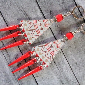 May include: A pair of unique earrings featuring a triangular design with a white background and a red and tan geometric pattern. The earrings have bright red, pointed accents and are attached to silver-colored hoops.