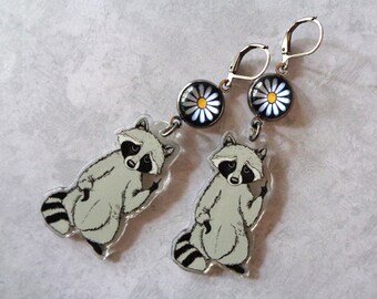 May include: A pair of silver earrings with a black and white raccoon charm and a white daisy charm. The raccoon charm is holding up its middle finger.