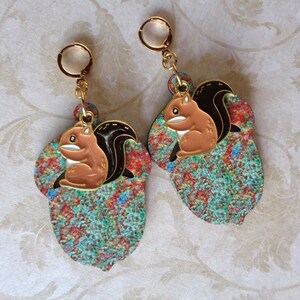 May include: A pair of gold-tone hoop earrings. Each earring features a colorful acorn shape with a brown squirrel charm. The squirrel has a black tail and is sitting inside the acorn. The acorn has a colorful pattern of red, blue, and green.