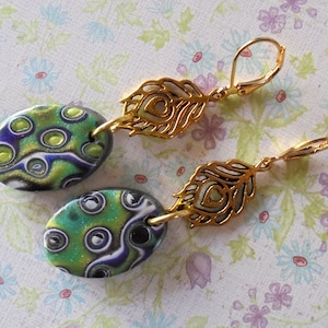 May include: A pair of earrings with green, white, and black oval-shaped polymer clay beads and gold-tone feather-shaped charms. The earrings have gold-tone leverback closures.