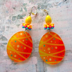 May include: A pair of orange and yellow Easter egg earrings with a beaded top. The earrings are made of clear plastic and have a swirl pattern.