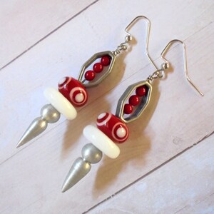 May include: A pair of dangle earrings with a unique design. Each earring features a silver-colored metal frame with three red beads, red and white patterned beads, white beads, and silver-colored cone-shaped accents. The earrings have silver-colored ear wires.