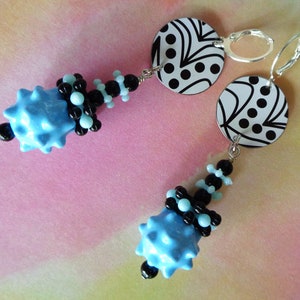 May include: A pair of dangle earrings featuring a blue and black bead cluster and a black and white patterned disc. The earrings are made with silver-toned metal.