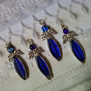 May include: Four angel-shaped pendants with silver-colored wings and accents. Each pendant features a long, oval, deep blue glass body and a small, round, blue bead at the top. The pendants are arranged on a white lace surface.