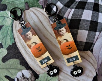 Pode incluir: Um par de brincos com tema de Halloween. Cada brinco apresenta um pingente retangular com uma imagem vintage de um bebê com um chapéu de bruxa segurando uma abóbora. A palavra "Boo" é impressa na parte inferior. Pingentes pretos pendem abaixo.