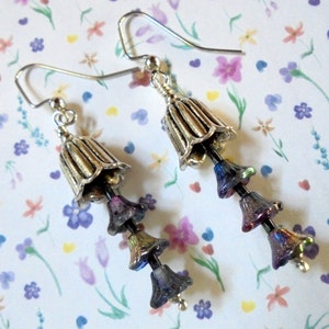 May include: Silver-toned bell-shaped earrings with iridescent purple and blue beads. The earrings have a floral design, with a bell-shaped top and three smaller bell-shaped beads hanging below.