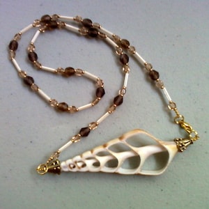 Puede incluir: Un collar artesanal con un colgante de concha natural. La cadena de cuentas es de color crema con detalles marrones y dorados. El colgante de concha es de color beige claro con intrincados calados.