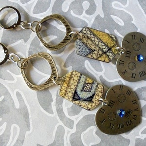 Puede incluir: Un par de pendientes colgantes con componentes de metal plateado. Cada pendiente presenta un cuadrado texturizado, una forma geométrica estampada en amarillo y azul, y un dije en forma de corazón con números romanos y una gema azul.