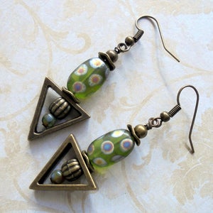 May include: A pair of handmade earrings featuring a triangular brass charm and a green glass bead with white and blue polka dots. The earrings have a vintage aesthetic with brass-colored metal accents and ear wires.