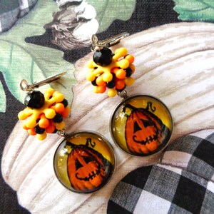 May include: A pair of Halloween-themed earrings. Each earring features a round, clear pendant with a smiling jack-o'-lantern design. The top of each earring has a cluster of yellow, orange, and black beads. The earrings are suspended from silver-colored hooks.