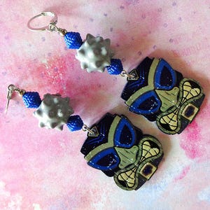 May include: A pair of dangle earrings featuring a tiki mask pendant in black, blue, and tan. Each earring includes a silver-toned metal leverback, a textured grey spherical bead, and two small, bright blue faceted beads connecting the components.