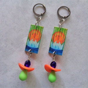 May include: A pair of dangle earrings. Each earring features a rectangular charm with a watercolor painting of a sunset over a lotus flower. Below the charm are orange and green shapes with a purple bead. The earrings are connected with silver-colored metal.