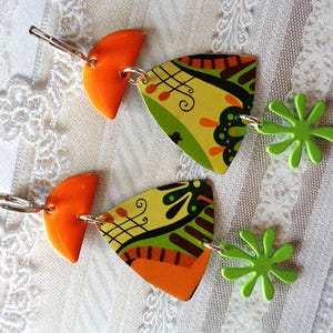 Puede incluir: Un par de pendientes únicos con diseños geométricos en naranja, amarillo, verde y negro. Los pendientes tienen una parte superior en forma de semicírculo naranja, un triángulo estampado y un dije de estrella verde. Se sujetan con ganchos plateados.