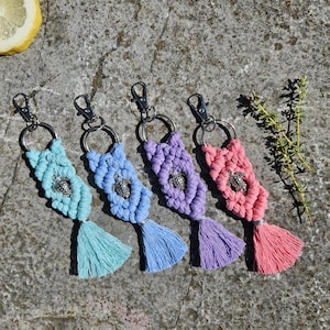 Puede incluir: Cuatro llaveros de macramé en tonos azul, morado y rosa, cada uno con un dije de tortuga plateada y una borla a juego. Los llaveros están sujetos a mosquetones y anillas de metal plateado.