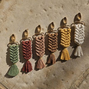 Puede incluir: Seis llaveros de macramé en varios colores: verde, marrón rojizo, beige, marrón, amarillo y blanquecino. Cada llavero presenta un diseño tejido, un cierre dorado y una borla a juego. Los llaveros están dispuestos en fila sobre una superficie texturizada.