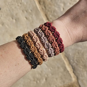 May include: A selection of macrame bracelets in shades of black, brown, beige, and burgundy. Each bracelet has a woven pattern and small gold beads. The bracelets are shown on a wrist.