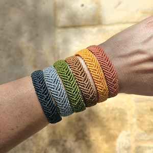 Peut inclure: Plusieurs bracelets tissés colorés, allant du bleu au orange, en passant par le gris, le vert, le marron et le jaune. Les bracelets, ornés d'un motif en chevrons, sont portés sur un bras. Chaque bracelet mesure environ 1,3 cm de large.
