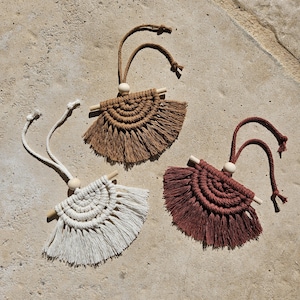 Può includere: Tre ornamenti a forma di ventaglio in macramè nei toni del marrone, bianco e bordeaux. Ognuno ha un tassello di legno e una perla di legno. Gli ornamenti sono appesi a un cordoncino ritorto. Lo sfondo è una superficie strutturata marrone chiaro.
