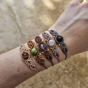 Puede incluir: Seis pulseras de macramé con detalles de piedras preciosas en varios colores, incluyendo marrón, verde, morado y negro. Cada pulsera presenta una piedra preciosa central y pequeñas cuentas doradas. Las pulseras se muestran en un brazo.
