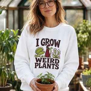 May include: A white long-sleeved sweatshirt featuring a graphic print with the text I GROW WEIRD PLANTS in dark purple letters. The design includes illustrations of a green Venus flytrap, a red pitcher plant, and a green sundew plant.