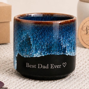 May include: A ceramic mug with a blue and black glaze, featuring the text "Best Dad Ever" in white. The mug has a brown rim and a heart symbol after the text. A gift tag that says "Happy Father's Day" is in the background.