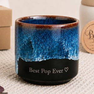 May include: A ceramic mug with a blue and black glaze, featuring the text "Best Pop Ever" and a heart symbol. The mug has a brown rim and is likely a Father's Day gift.