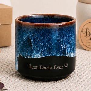 May include: A ceramic mug with a blue and black glaze, featuring the text "Best Dada Ever" and a heart symbol. The mug has a brown rim and is likely a Father's Day gift. A gift box and tag that says "Happy Father's Day" are in the background.