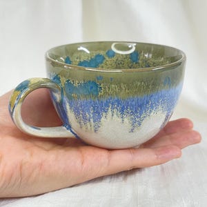 May include: A handmade ceramic coffee cup with a rounded shape and a handle. The cup features a color gradient, with a white base, transitioning to blue, then green at the rim. The handle has yellow and blue accents.