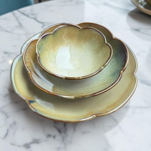 May include: A trio of ceramic serving dishes with a scalloped edge. The set includes a large plate, a medium plate, and a small bowl. The dishes have a gradient colour scheme, transitioning from light yellow to a darker green, with a brown rim.