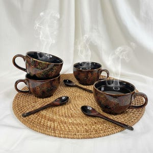 May include: A set of four ceramic coffee cups with matching spoons. The cups are dark brown with a speckled glaze, and the spoons are a similar color. The cups are arranged on a round woven placemat.