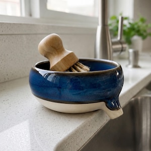 May include: A blue and white ceramic dish with a wooden handled brush. The dish has a spout and is sitting on a white countertop next to a stainless steel sink. The brush is made of wood and bristles.