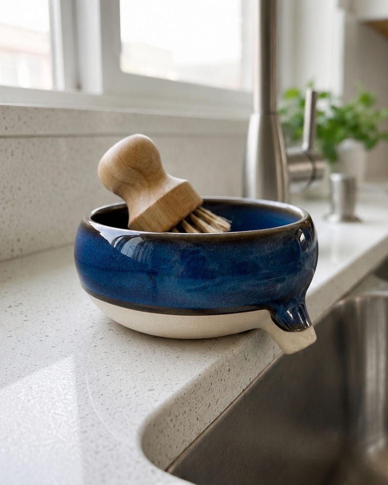 May include: A blue and white ceramic dish with a wooden handled brush. The dish has a spout and is sitting on a white countertop next to a stainless steel sink. The brush is made of wood and bristles.