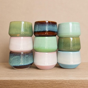 May include: A collection of nine small, ceramic bowls in various colors, stacked in three rows. The bowls feature a rounded shape and a glossy finish. Colors include green, blue, pink, and white, with some bowls showcasing a two-tone design.